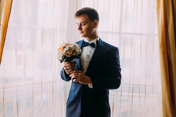 Elegante vestido noivo romântico segurando um buquê de casamento de rosas rosa pálidas em mãos de pé perto da janela — Fotografia de Stock