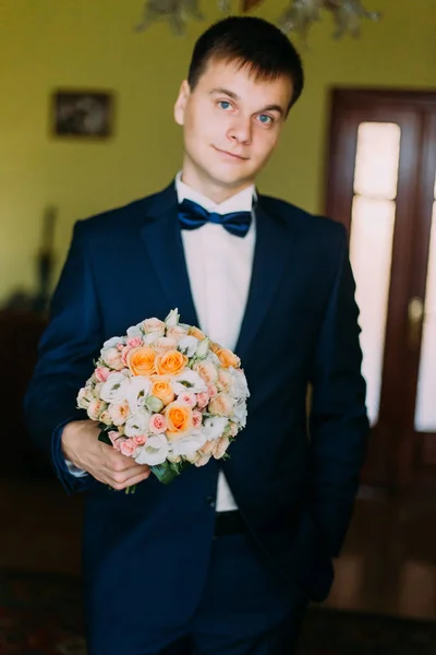 Ellegant klädd eleganta brudgummen håller en bröllop bukett vit och blek orange rosor i händer stående i hotellrum — Stockfoto