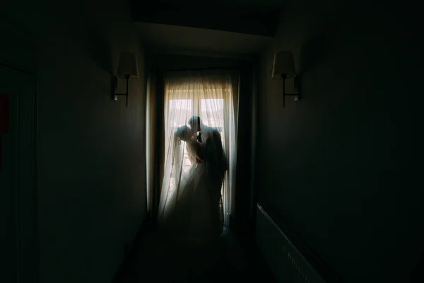Öpüşme yeni evli çift ayakta pencerenin karanlık odada yakınındaki silüeti. — Stok fotoğraf