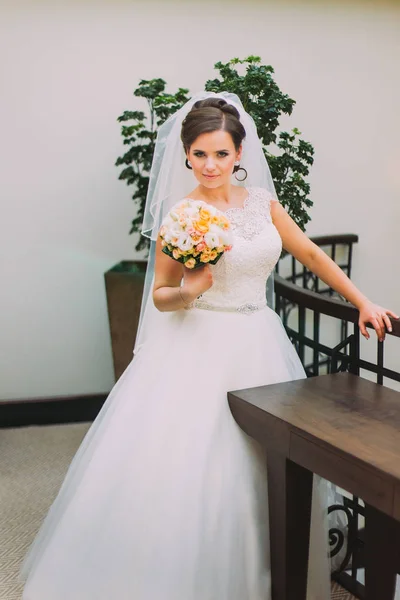 Hermosa novia en vestido blanco de pie cerca del balaustrer celebración de ramo de boda de rosas rosadas pálidas — Foto de Stock