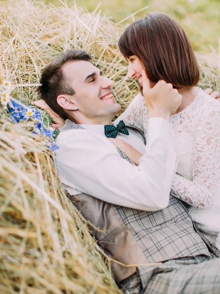 Retrato sensible de los recién casados sonrientes. El novio está acariciando la mejilla de la novia mientras está acostado en el heno . — Foto de Stock