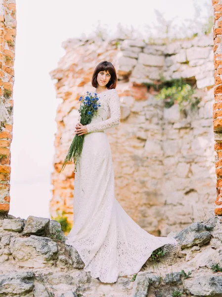 La bella sposa sta tenendo il bouquet con fiori selvatici blu mentre in piedi sui suini della casa . — Foto Stock