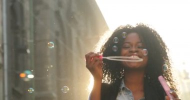 Güneş ışınları arka plan sabun köpüğü üfleme gülümseyen Afro-Amerikan kız portresi.