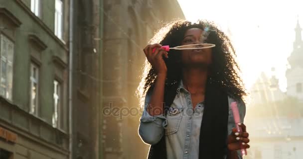 Mulher afro-americana energética com cabelo encaracolado está soprando as bolhas de sabão na rua durante o dia ensolarado . — Vídeo de Stock