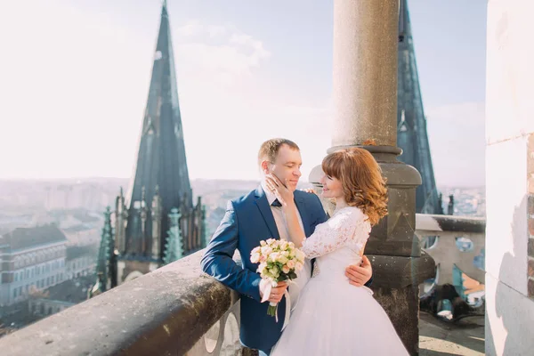 Newlywed bride and groom posing on the balcony of old gothic cathedral with city view at background — Stock Photo, Image