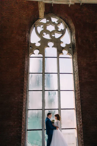 Happy stylish bride and groom holding each other in front of arched window old gothic cathedral — Stock Photo, Image