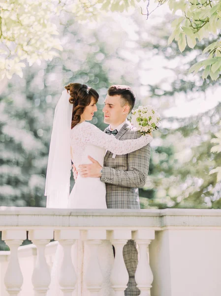 La foto vertical de los recién casados felices abrazándose detrás de los barriles de la vieja casa . — Foto de Stock