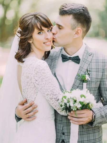 El novio con el ramo de la boda está besando a la novia alegre en la mejilla . — Foto de Stock