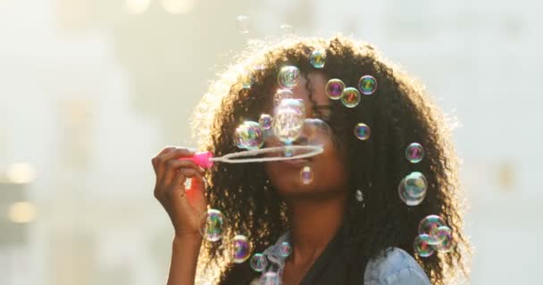 Retrato de cerca de la hermosa sonriente afro-americana con el pelo rizado soplando burbujas de jabón en la calle . — Vídeos de Stock