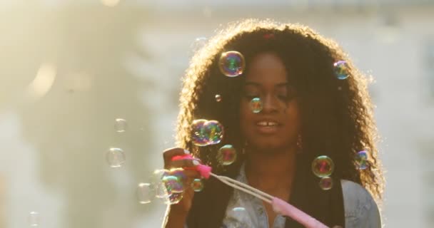 Retrato horizontal de la encantadora afro-americana con el pelo rizado soplando las burbujas de jabón en el fondo de los rayos del sol . — Vídeos de Stock