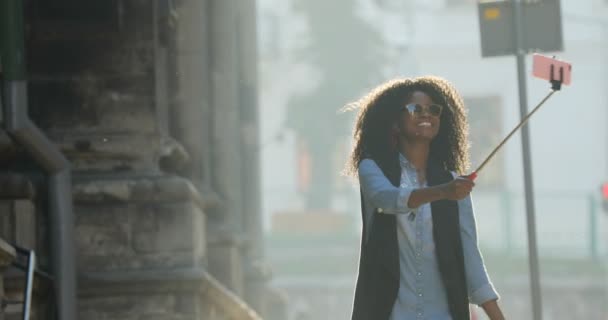 Nádherný afro-american model s kudrnatými vlasy nosí sluneční brýle se ukazuje znamení míru při pořizování fotografií pomocí selfie stick během chůze po ulici. — Stock video