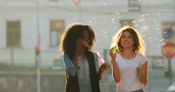 Veselá přítelkyně vane mýdlové bubliny při chůzi po ulici. Jedním z nich je afro-american. — Stock video