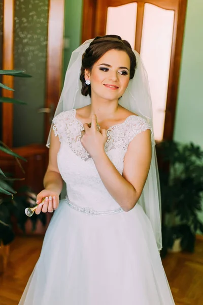 Encantadora novia en vestido blanco magnífico usando perfume antes de la ceremonia de boda —  Fotos de Stock
