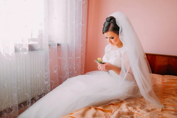 Bela noiva sentado em uma cama perto da janela em espera e segurando bonito pouco casamento boutonniere — Fotografia de Stock