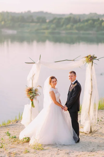 Glada nygift par håller händerna under deras bröllopsceremoni nära floden. — Stockfoto