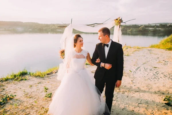 La vista horizontal de los recién casados felices caminando brazo en brazo cerca del río . — Foto de Stock