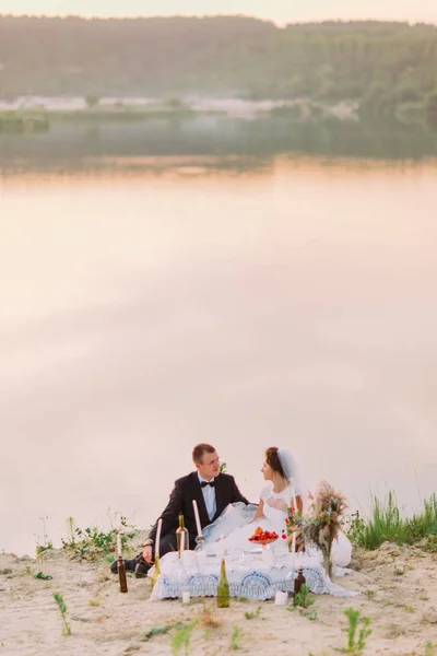 Das ganzseitige Foto des Brautpaares beim Hochzeitspicknick im Hintergrund des Flusses während des Sonnenuntergangs. — Stockfoto