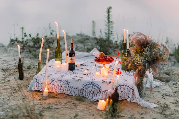 El romántico juego de picnic decorado con velas brillantes en las botellas, fruta y ramo de flores silvestres . —  Fotos de Stock