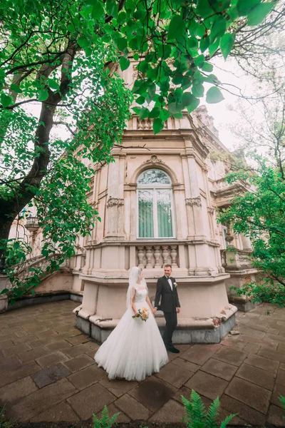 La foto vertical de los recién casados caminando cerca del antiguo edificio rodeado de árboles . —  Fotos de Stock