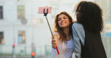 Güneş ışınlarının arka plan, sokakta yürürken selfie stick kullanarak fotoğraf çekmek sevimli gülümseyen kız sarılıyorlar. Yakın çekim görünümü.
