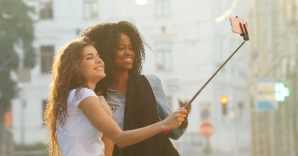 Horizontal side view of the beautiful smiling multiethnic girlfriends taking photos using the selfie stick in the street. — Stock Video