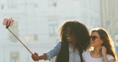 Yürüyüş sırasında selfie stick kullanarak fotoğraf çekerken hava öpücük gönderme iki neşeli çok ırklı kız arkadaşım Slow motion video.