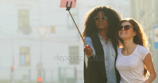 Joyful walk of two smiling multiethnic girlfriends with sunglasses taking photos using the selfie stick. — Stock Video