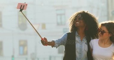 Güneş gözlüğü takıyor güzel gülümseyen kız güneşli Caddesi boyunca neşeli yürüyüş sırasında selfie stick kullanarak çekim.