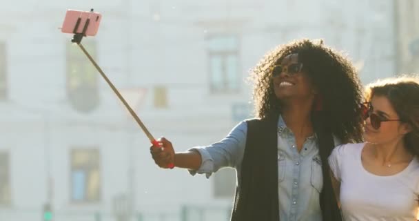 Belas namoradas sorridentes usando óculos de sol estão tirando fotos usando o pau de selfie durante o alegre passeio ao longo da rua ensolarada . — Vídeo de Stock