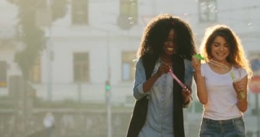 Gülümseyen Afro-Amerikan ve sarışın kız güneşli cadde boyunca yürürken sabun köpüğü üfleme vardır.