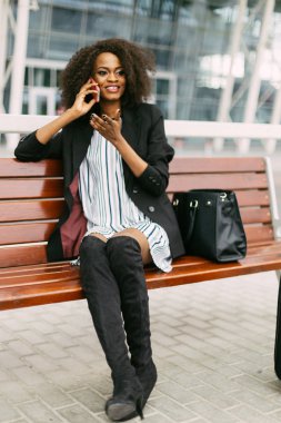 Şık giyimli gülümseyen Afro-Amerikan Gezgin bankta otururken cep telefonu aracılığıyla konuşuyor.