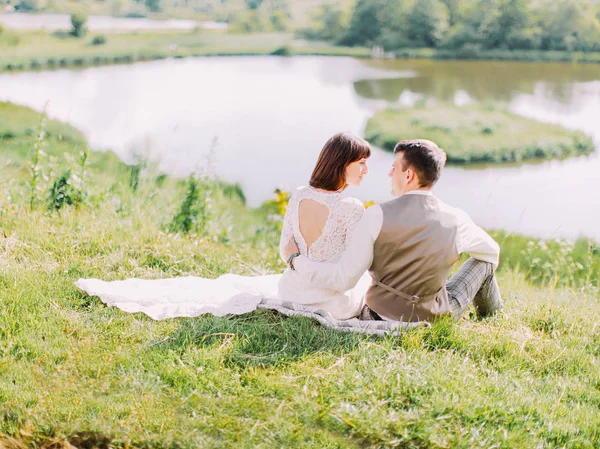 Sensível vista traseira ao ar livre dos recém-casados abraçando sentado no prado e olhando um para o outro . — Fotografia de Stock