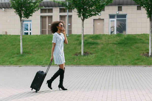 Vista orizzontale della allegra ragazza afro-americana che tiene i bagagli mentre parla tramite il telefono cellulare . — Foto Stock