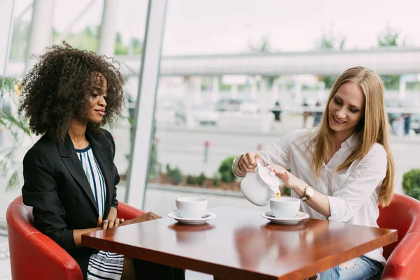 De mooie vrouw is het gieten van de thee tijdens de vergadering met de Afro-Amerikaanse vriendin. Locatie Cafe. — Stockfoto