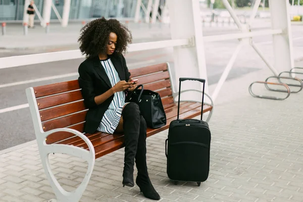 La bella donna afro-americana è seduta sulla panchina e chatta tramite il cellulare vicino all'aeroporto . — Foto Stock