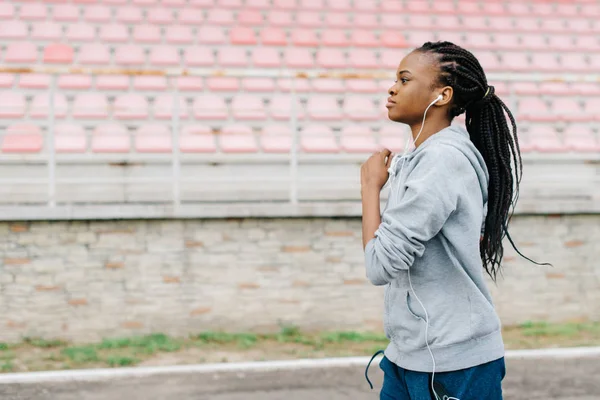 Side portrait of the athletic afro-american girl jogging along the stadium while listening to music in earphones.