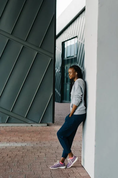 Kant full-length foto van de charmante fitness tiener in de oortelefoons leunend op de muur. Het meisje is de Afro-Amerikaans. — Stockfoto