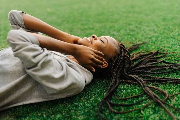 Primo piano ritratto laterale dell'affascinante ragazza afro-americana sorridente che si gode la musica negli auricolari mentre si sdraia sull'erba . — Foto Stock