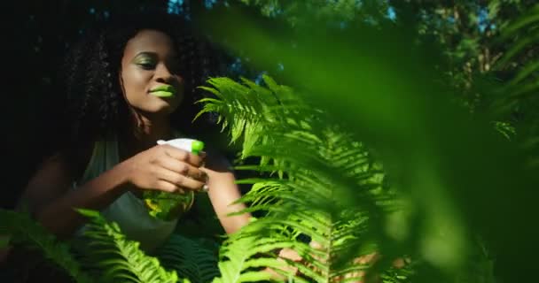 Das Outdoor-Porträt der charmanten Afroamerikanerin mit leuchtend grünem Make-up, die sich um den Farn kümmert. das junge Mädchen sprüht die Pflanzen mit dem grünen Pulverisierer. — Stockvideo