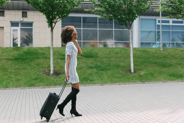 Vista laterale della donna afro-americana vestita con stile che tiene i bagagli mentre parla via cellulare . — Foto Stock