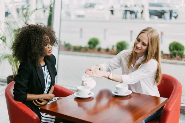 The meeting of two beautiful women. The afro-american is talking while the bolnde one is pouring the tea in the cafe. — Stock Photo, Image