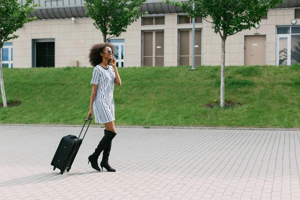 Mulher afro-americana bonita está falando através do telefone celular enquanto segurando a bagagem . — Fotografia de Stock