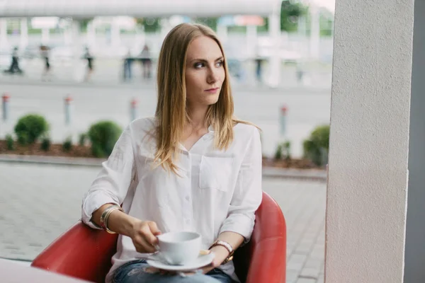 Horizontální portrét blond ženy dívá stranou, zatímco držíte šálek kávy v kavárně. — Stock fotografie