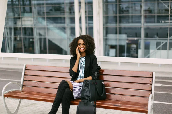Крупный план портрета красивой улыбающейся афро-американской путешественницы, разговаривающей по мобильному телефону в ожидании равнины на скамейке . — стоковое фото