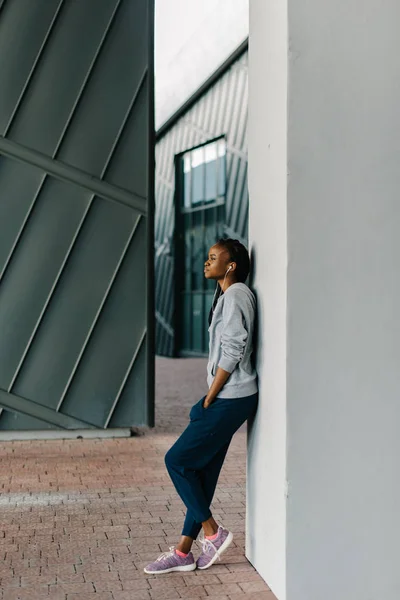 De charmante Afro-Amerikaanse sportwoman luistert naar muziek in oortelefoons terwijl leunend op de muur. De full-length zijaanzicht. — Stockfoto