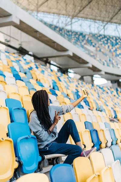 A vista de trás do belo adolescente afro-americano mostrando sinal de paz ao tirar selfie nos assentos do estádio . — Fotografia de Stock