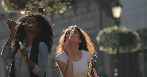 The close-up view of the beautiful cheerful multiculticultural girlfriends blowing the soap bubbles during the walk in the street. — Stock Video