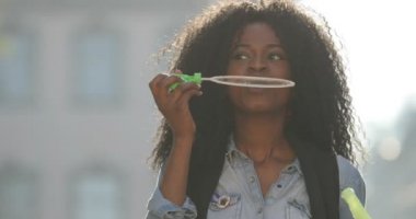 Kıvırcık saçlı iplik güneşli sokakta sabun köpüğü üfleme sırasında yuvarlak ile muhteşem Afro-Amerikan kız portresi.