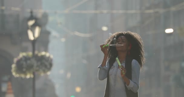 A encantadora menina afro-americana alegre está soprando bolhas de sabão enquanto balança ao redor da rua ensolarada . — Vídeo de Stock