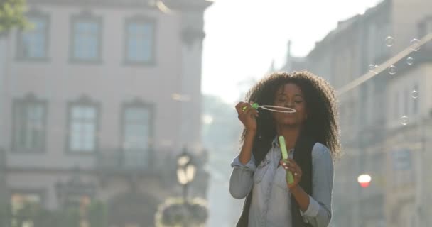 Porträtt av leende stilfullt klädd afro-american flickan blåser såpbubblor och snurrar runt på gatan. — Stockvideo